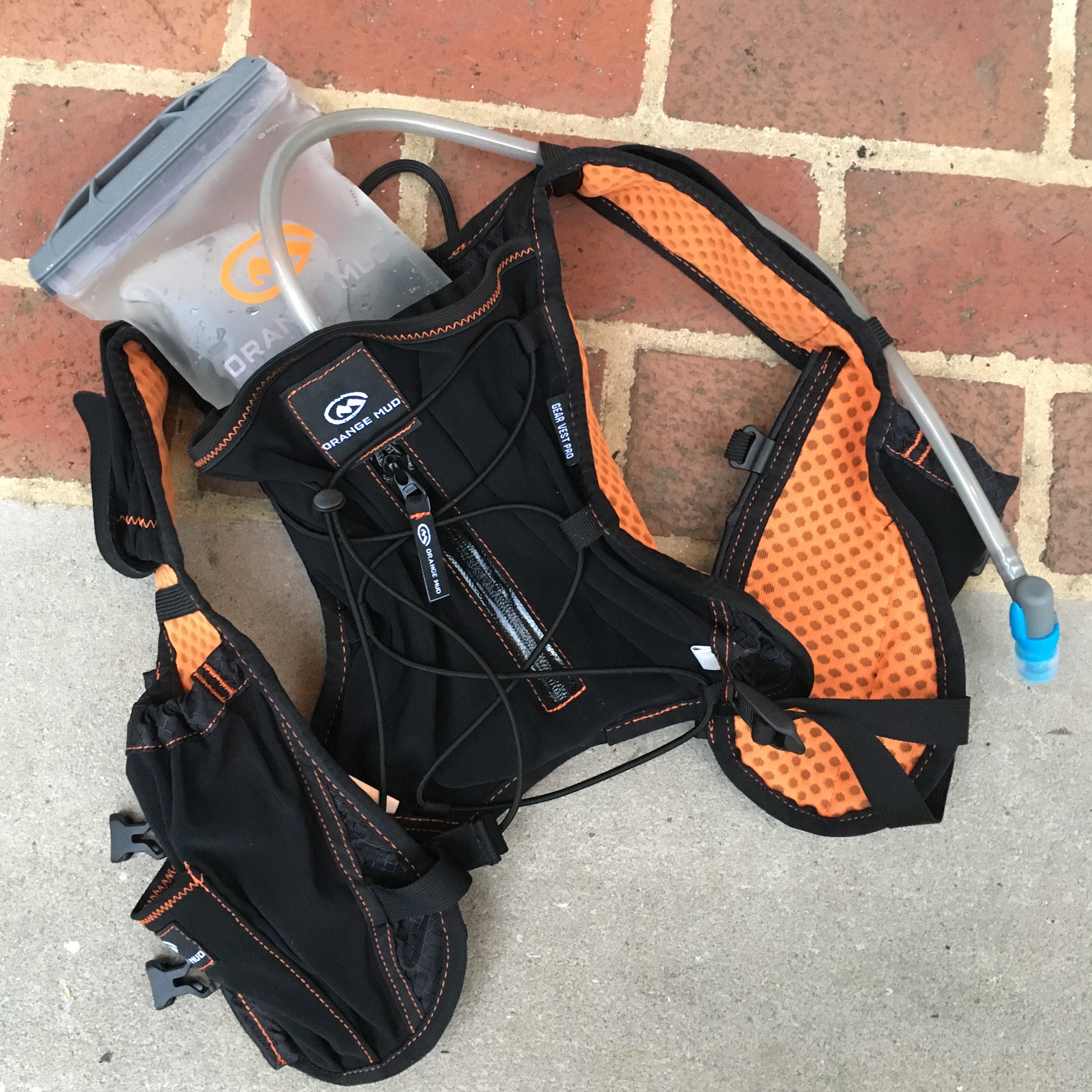 Orange Mud Gear Vest Pro, showing bladder holding water, against brick/concrete background. 