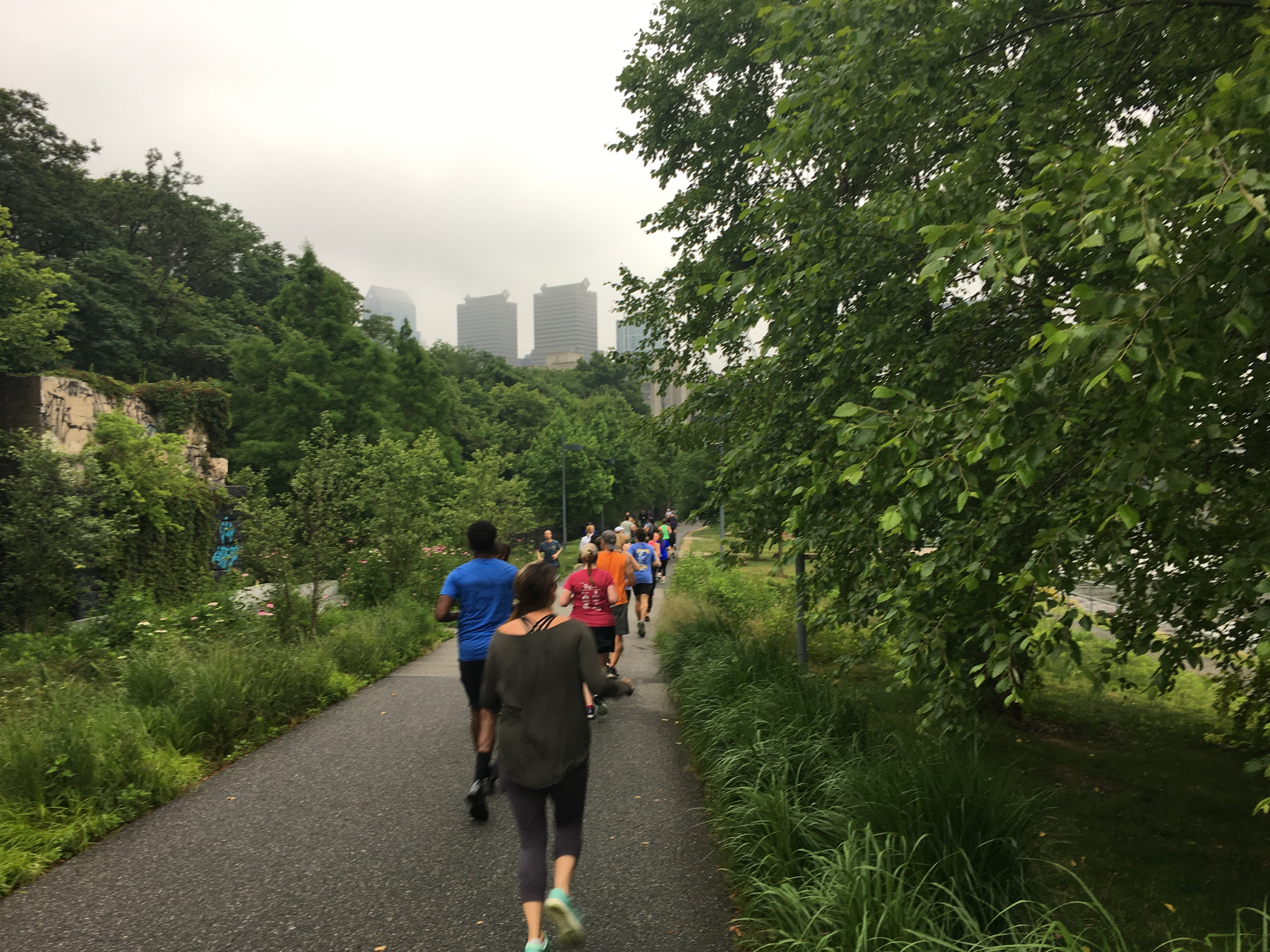 Runners on Schuylkill River Trail with some skyscrapers in the distance and trees on both sides. 