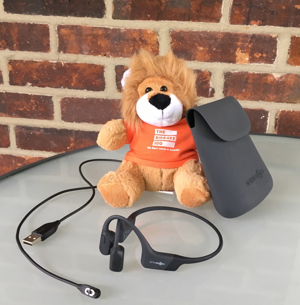 Benjamin, The BibRave 100 mascot, posed with the AfterShokz Aeropex, charging cord and case against a brick background. 