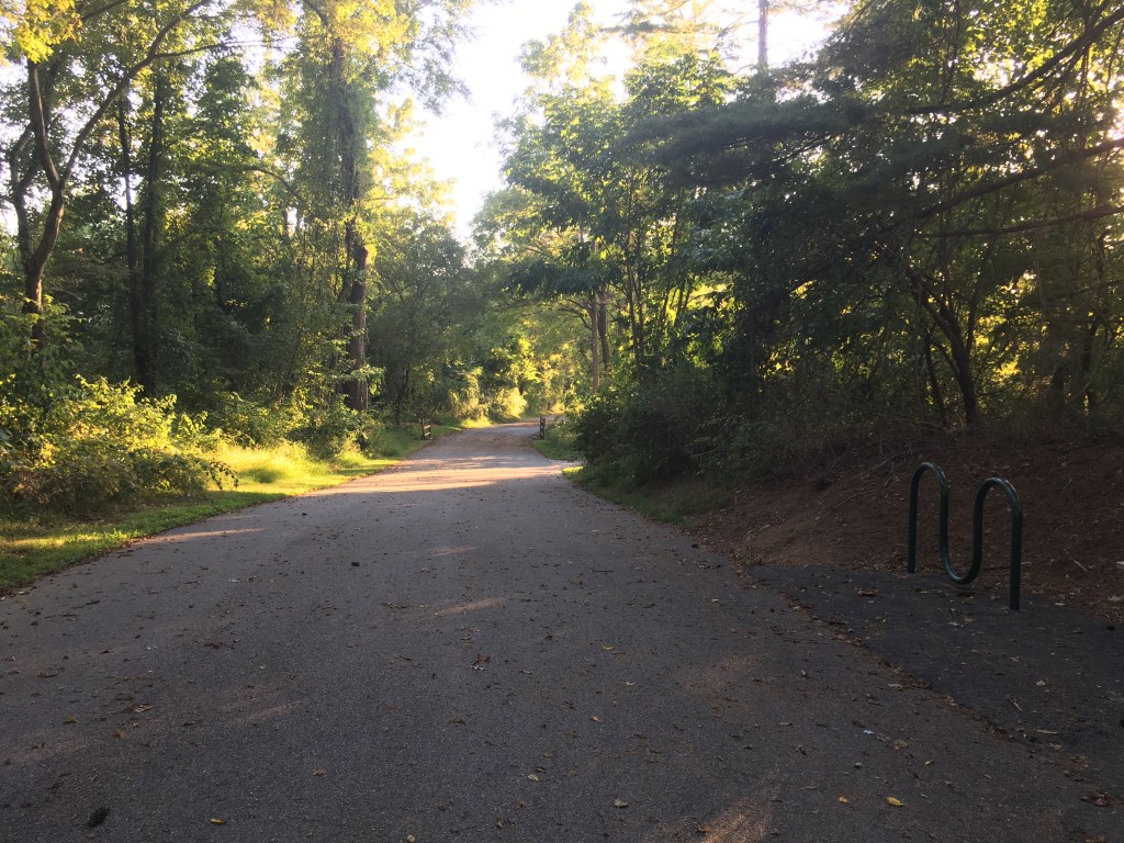 View of paved trail with trees on both sides and a bike rack (with no bikes on it) to the right. 
