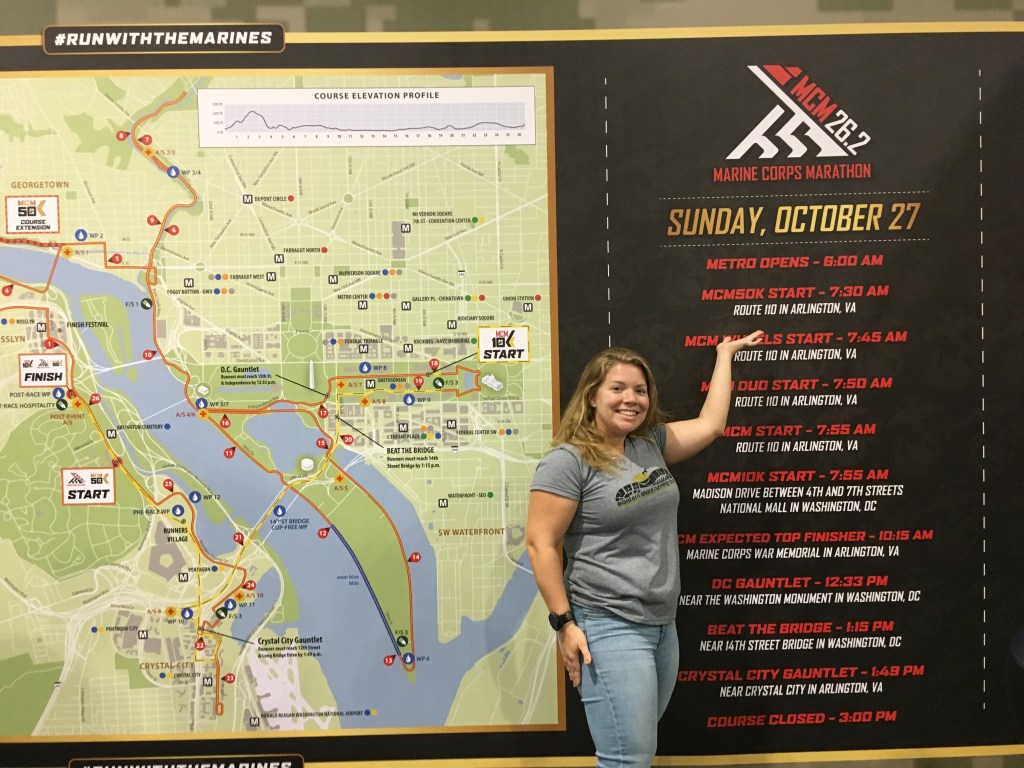 Vanessa Junkin standing next to a map of the Marine Corps Marathon course and pointing to her birthday, which is above the schedule, on a display/sign.