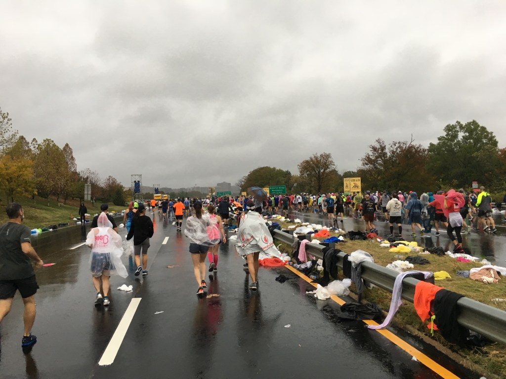 Runners walk on the highway ahead of the Marine Corps Marathon, with a cloudy sky overhead.
