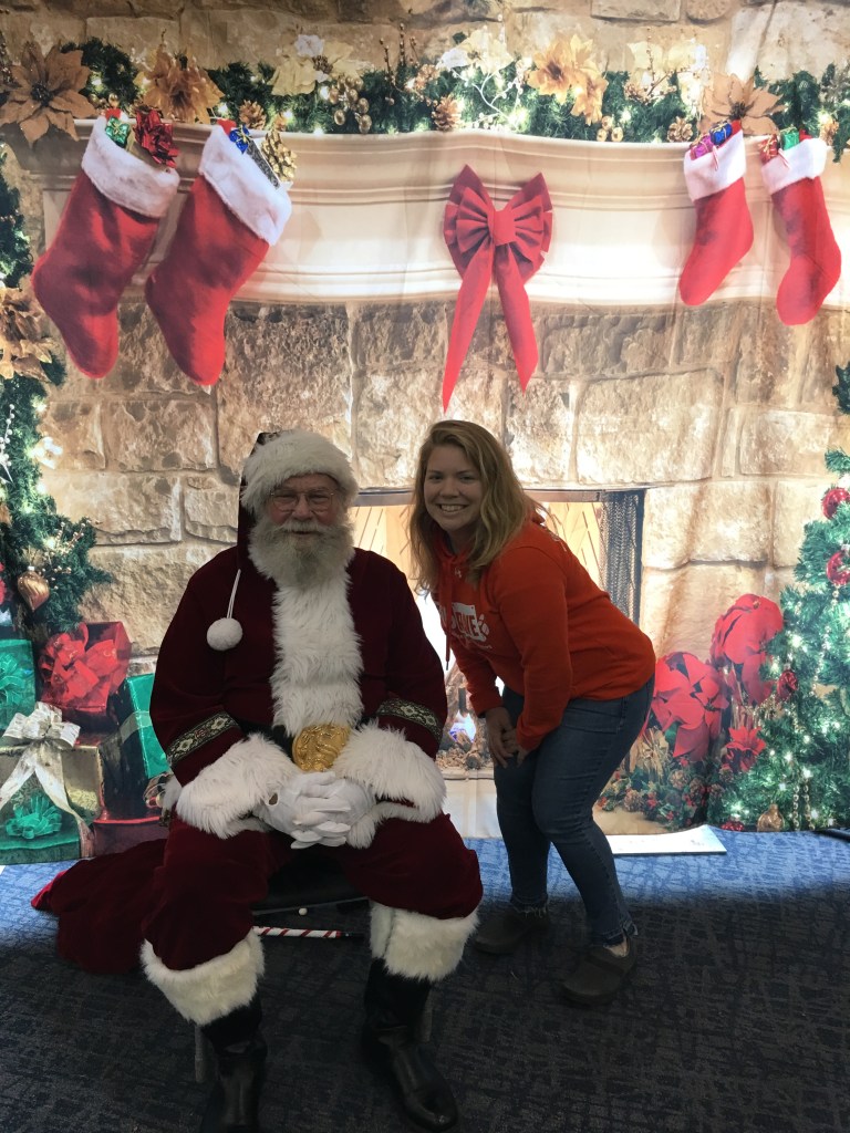 Vanessa Junkin crouching next to a sitting Santa Claus against a Christmas chimney backdrop.