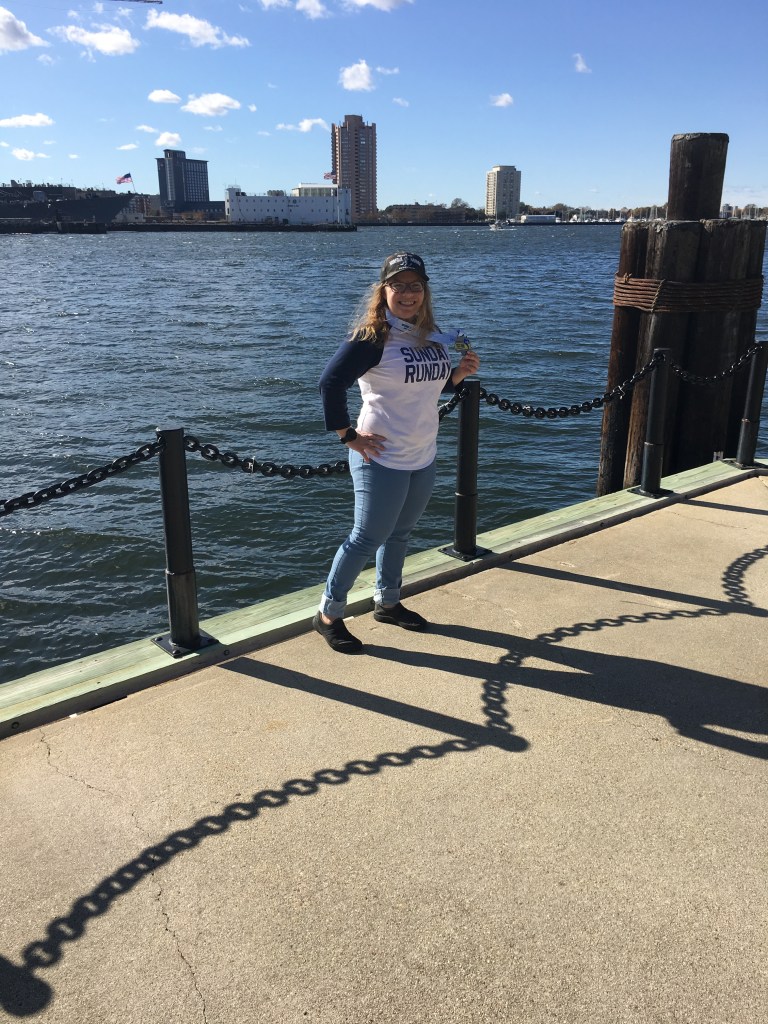 Vanessa posing against the waterfront with her medal, wearing a "Sunday Runday" shirt and the finisher hat.