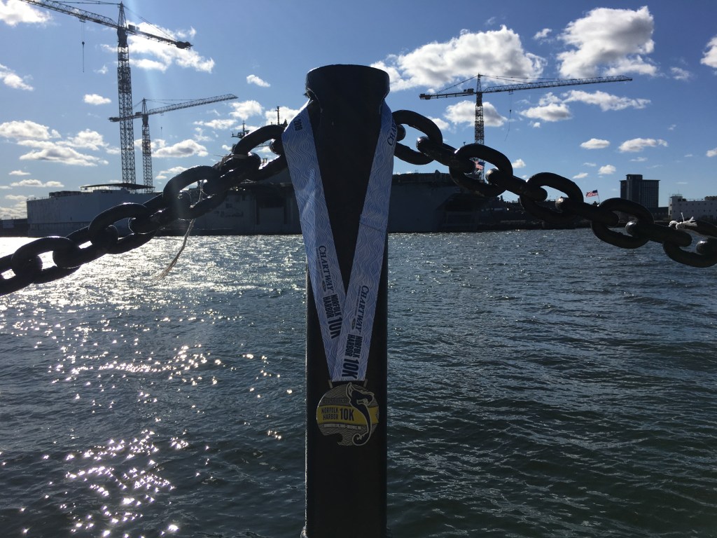 Norfolk Harbor 10K medal shown hanging on a metal pole in front of the waterfront and a ship.