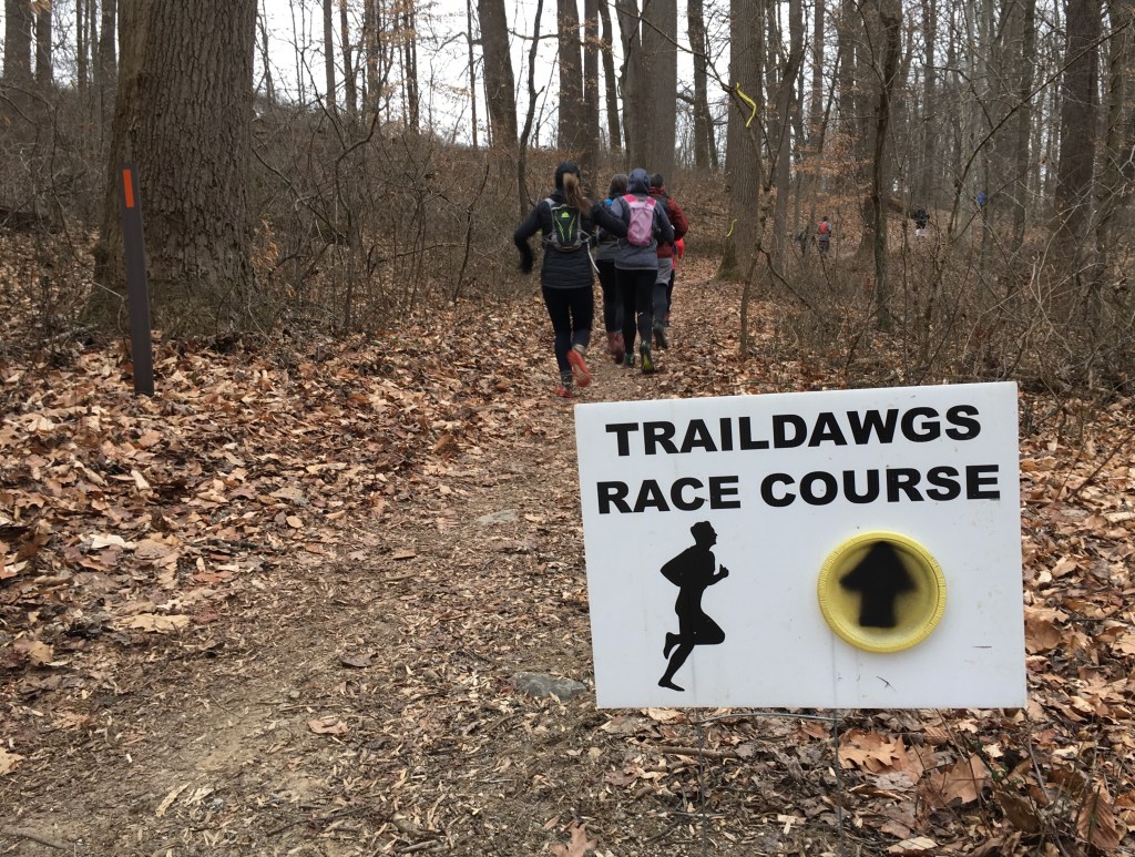 "Traildawgs Race Course" sign in the foreground with runners on a trail behind it. 