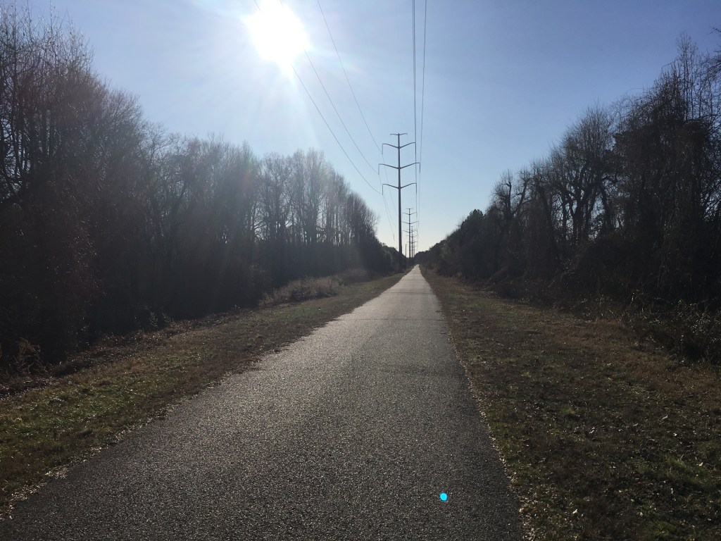 Paved trail is shown with grass and trees on both sides and power lines along the trail. 