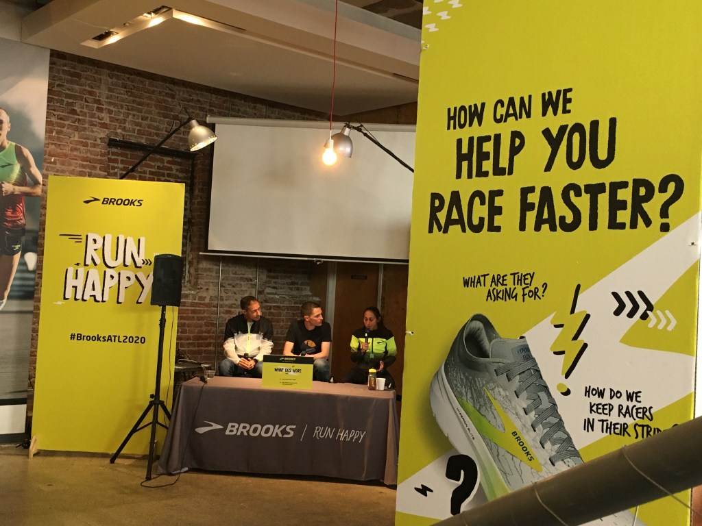 Three people at a Brooks table; pro runner Des Linden is speaking. Brooks displays are shown. 
