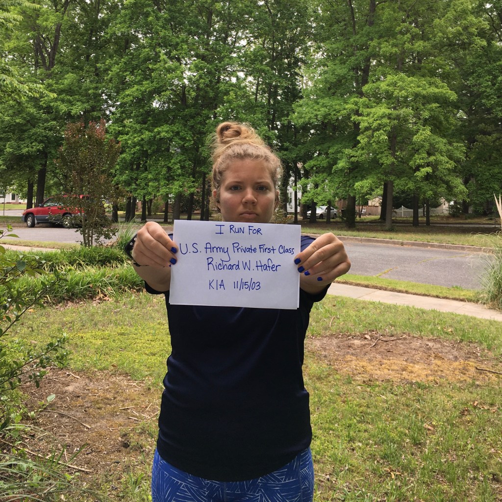 Vanessa Junkin holding out a piece of paper that says "I run for U.S. Army Private First Class Richard W. Hafer KIA 11/15/03." 