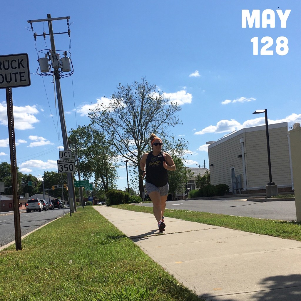 Woman running on a sidewalk with a building with siding on the right side and a road to the left.