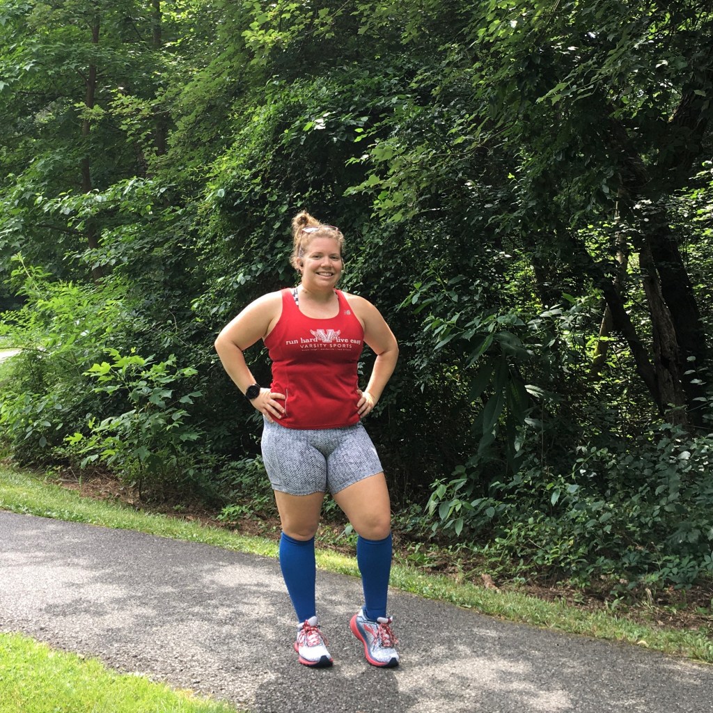 Vanessa Junkin, a female runner, poses with her hands on her hips in a red tank top, gray herringbone shorts and long blue socks. 