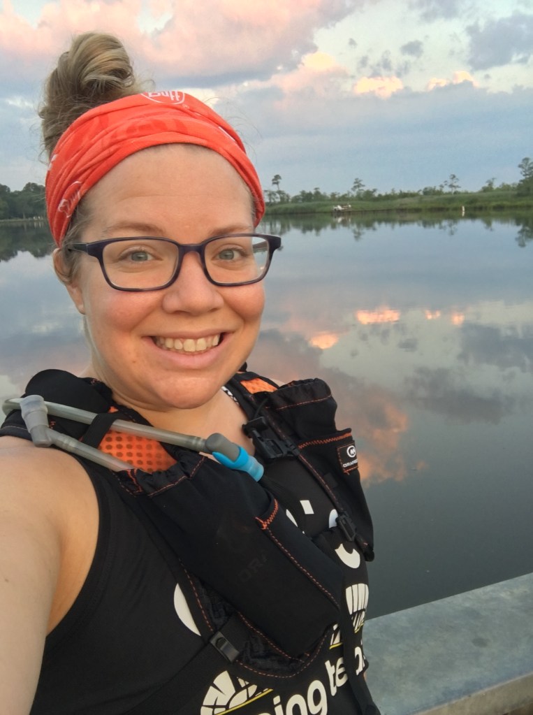 Selfie of Vanessa Junkin, wearing hydration pack and orange buff, in front of the water and sunrise. 