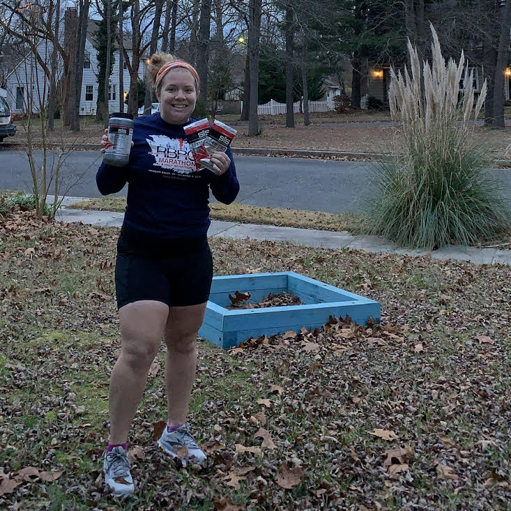 Female runner stands in front yard holding canister of Science in Sport REGO Rapid Recovery in one hand and two packets in the other.
