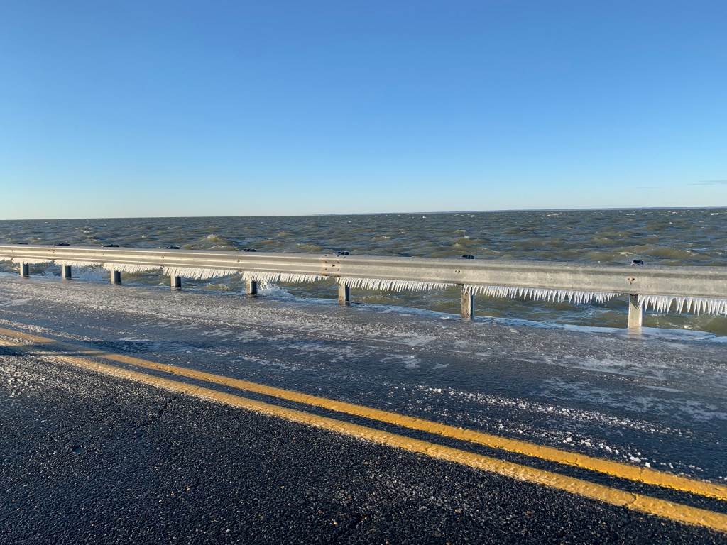 A road next to choppy water, with icicles formed on the guardrail. 