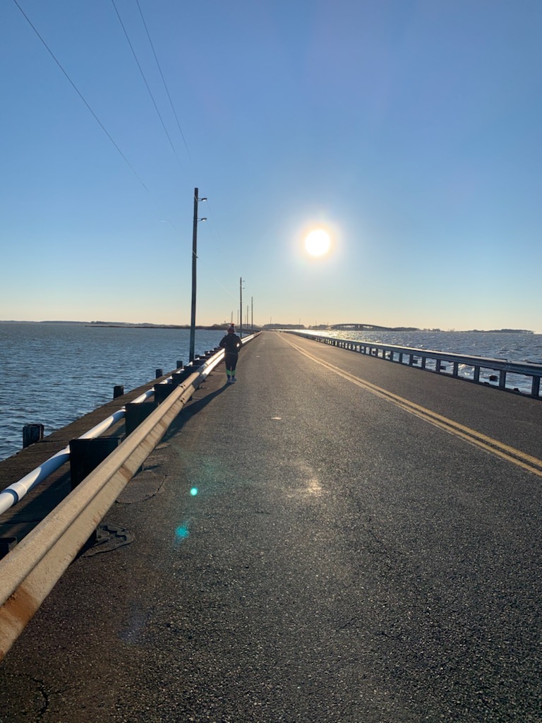 Runner in distance running on road with water on both sides. 