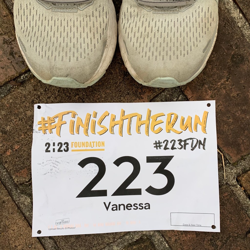 The tops of running shoes above a race bib on the ground that reads #FinishTheRun, 2:23 Foundation and #223FDN, with the number "223" and "Vanessa" under that. 
