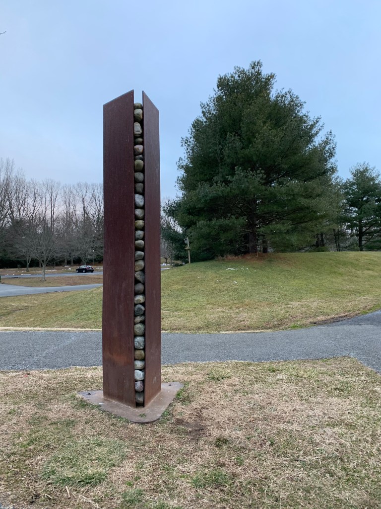 Sculpture that has what looks like tall metal rectangles going up, with rocks in the middle. A paved trail is behind it. 