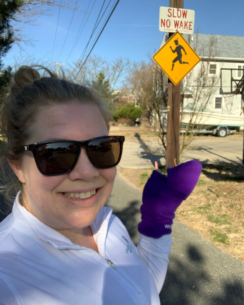 Vanessa Junkin, wearing purple WhitePaws RunMitts, gestures to a sign that says "Slow No Wake" with a runner in a yellow diamond sign underneath. 