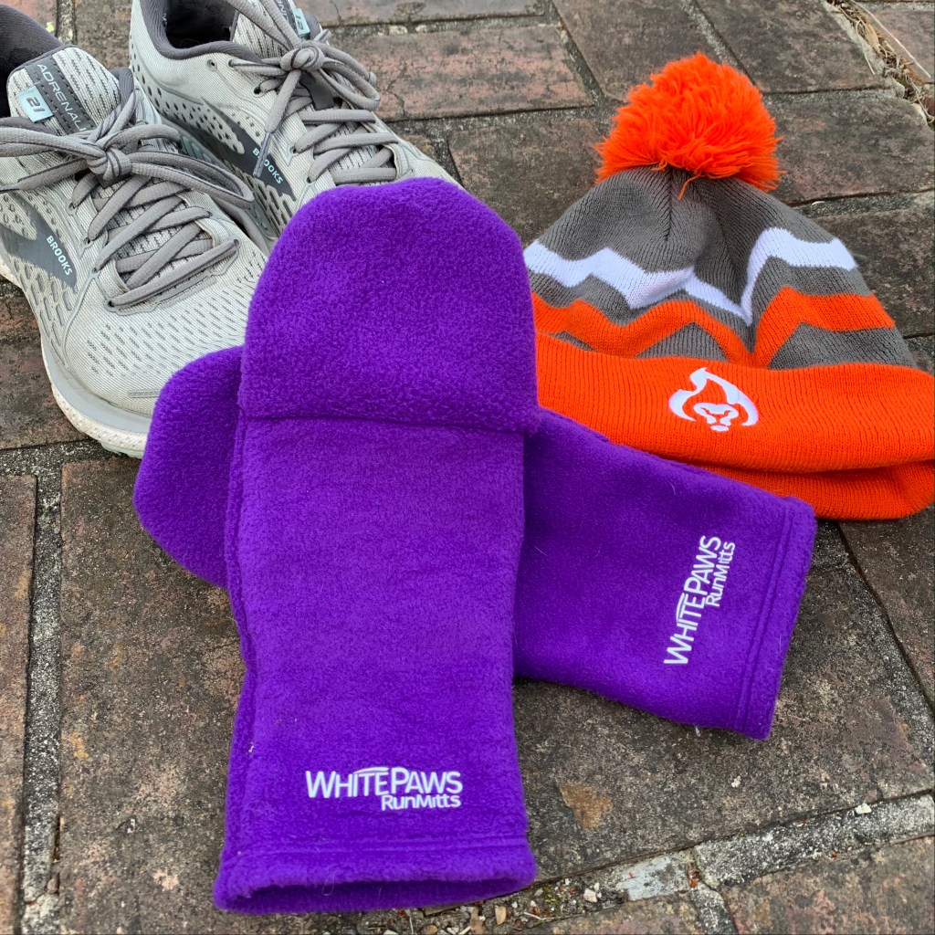 A pair of purple WhitePaws RunMitts are shown with a pair of Brooks Adrenaline running shoes to the left and an orange and gray BibRave winter hat to the right. 