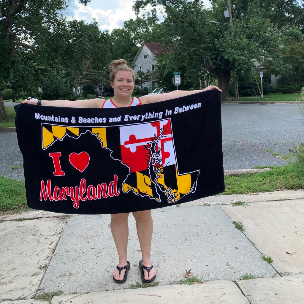 Vanessa Junkin holds up black towel that has a shape of Maryland with the Maryland flag design. It says "Mountains & Beaches and Everything in Between" at the top and "I heart Maryland."