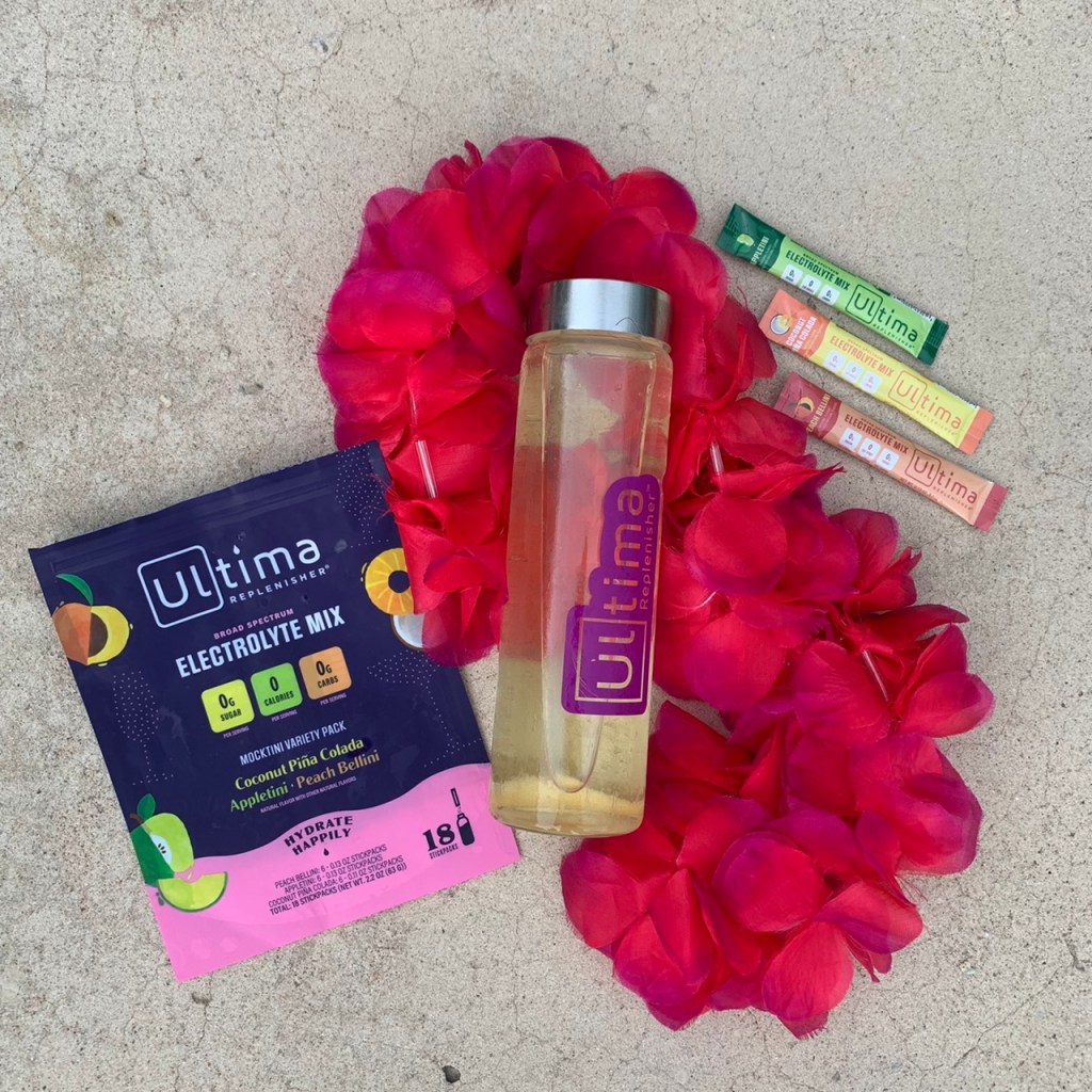 Ultima Replenisher package, filled glass water bottle on top of a pink Hawaiian lei and three stickpacks of Ultima Replenisher flavors.
