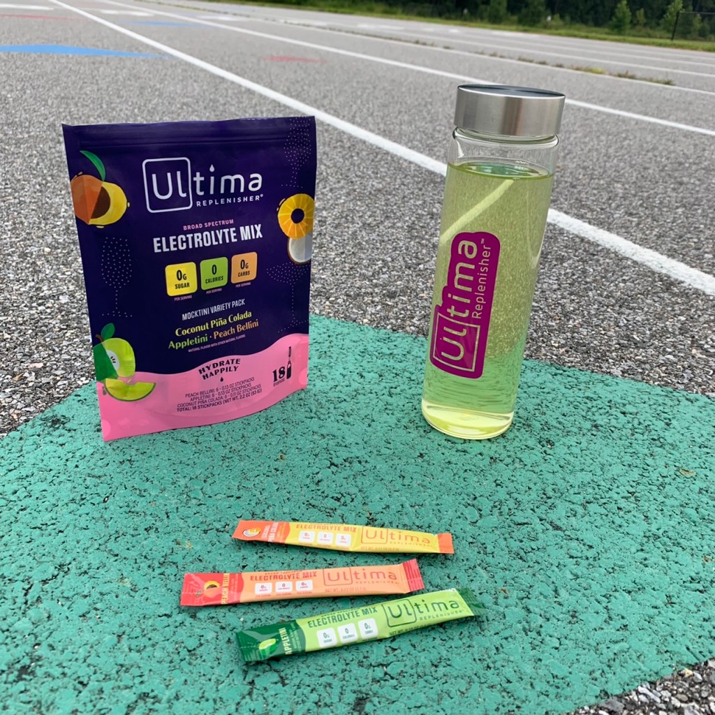 Ultima Replenisher Mocktini Variety Pack package on top of running track next to a glass bottle with green liquid in it. Three stickpacks are in the front.