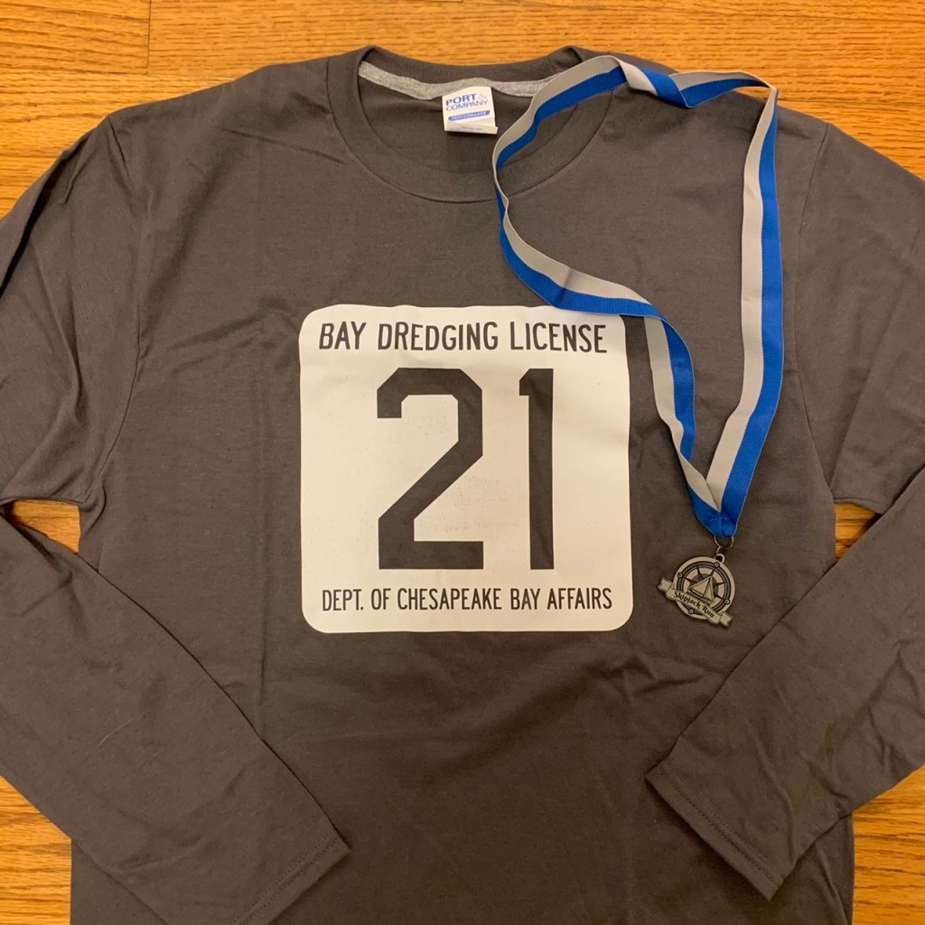 Gray long-sleeved shirt with a white rounded square that says "Bay Dredging License / 21 in large font / Dept of Chesapeake Bay Affairs," along with small medal. 