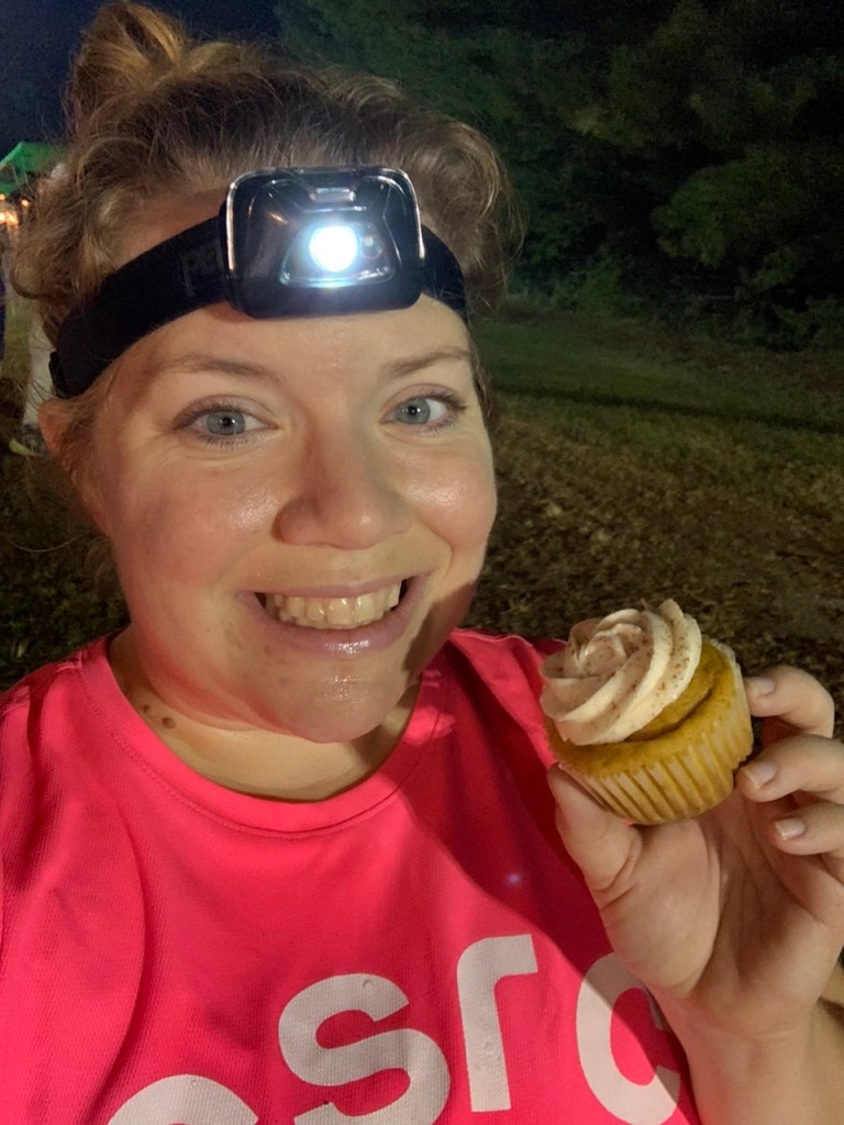 Vanessa Junkin, wearing a pink shirt and a headlamp, takes a selfie while holding a cupcake. 
