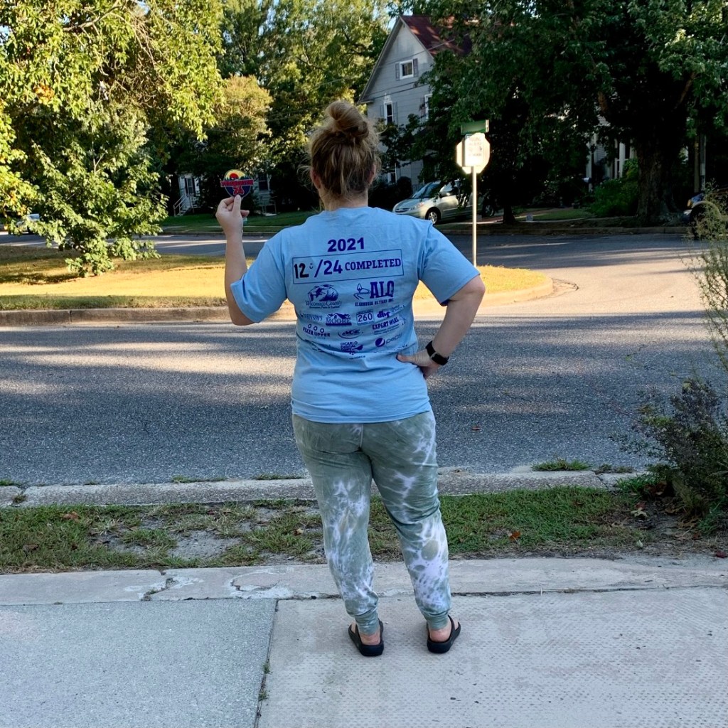 Back of Vanessa Junkin standing up with "12/24 completed" showing on shirt. She is also holding up her patch. 