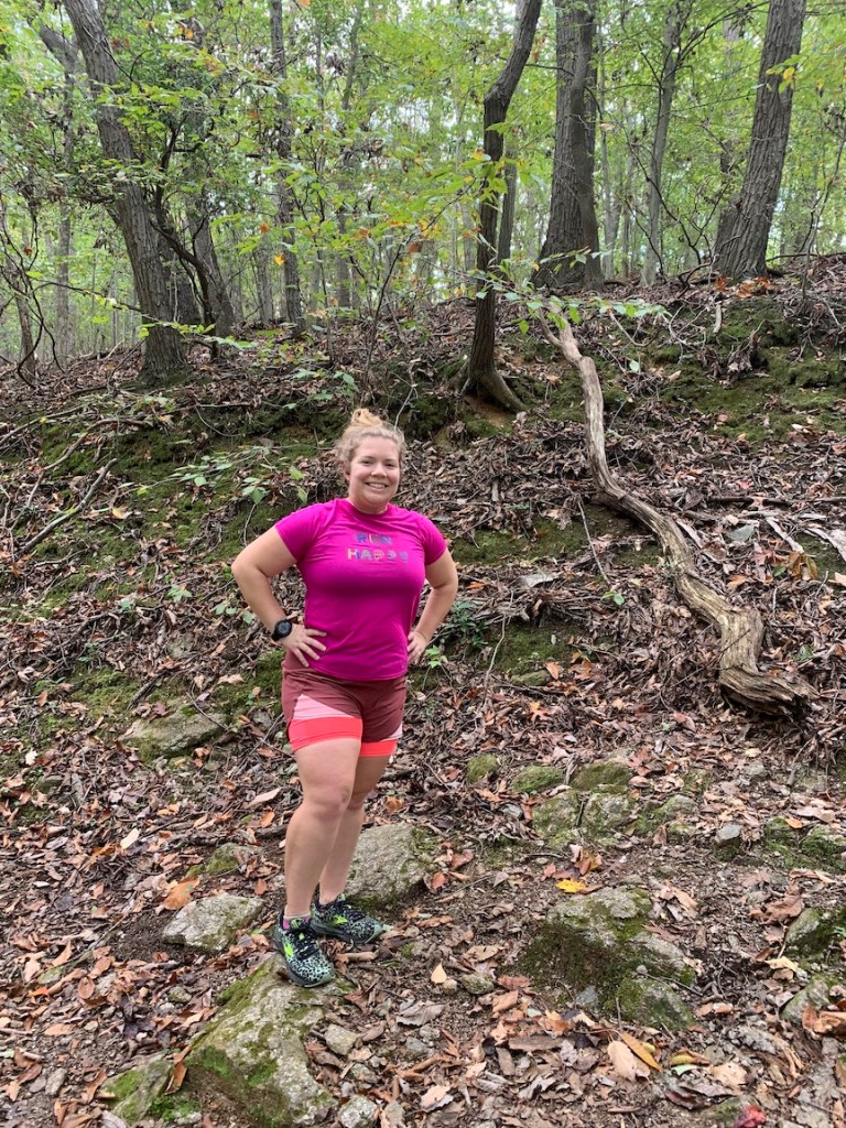 Vanessa Junkin poses in running clothes and shoes by a rocky trail in the woods. 