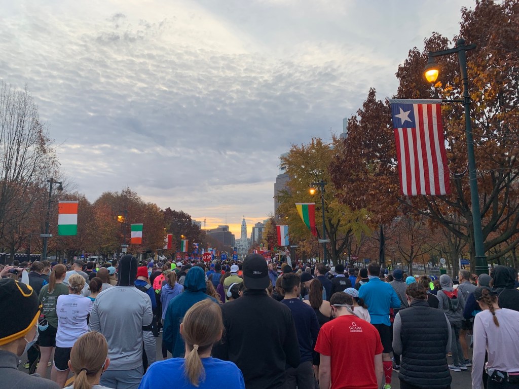 Runners wait to begin the Philadelphia Marathon. There are country flags on both sides and skyscrapers in the distance.