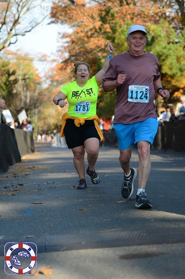 Vanessa Junkin puts her arm up holding a pacing stick while running, about to finish the race, with a happy runner in front.  