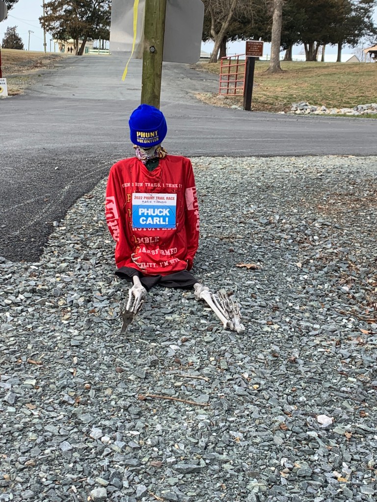 Skeleton sitting on the ground with PHUNT hat, shirt and a bib reading "PHUCK CARL." 