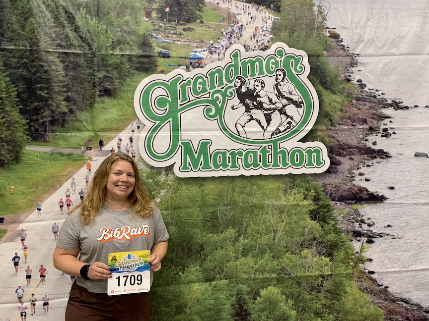 Vanessa Junkin holds race bib and poses in front of a Grandma's Marathon backdrop. 