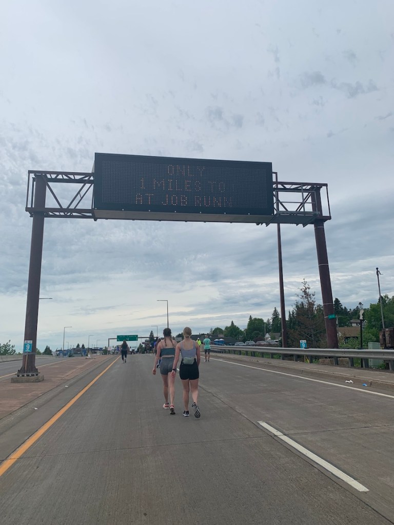 Road sign reads "Only 4.1 Miles to Go Great Job Runners" as race participants make their way up a hill. 