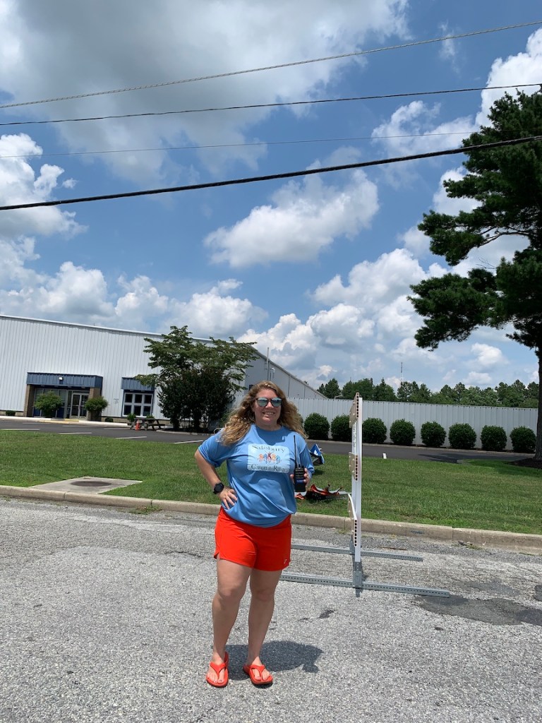 Vanessa Junkin poses in a sunglasses, a light blue shirt and dark orange shorts, while holding a walkie talkie outside. 
