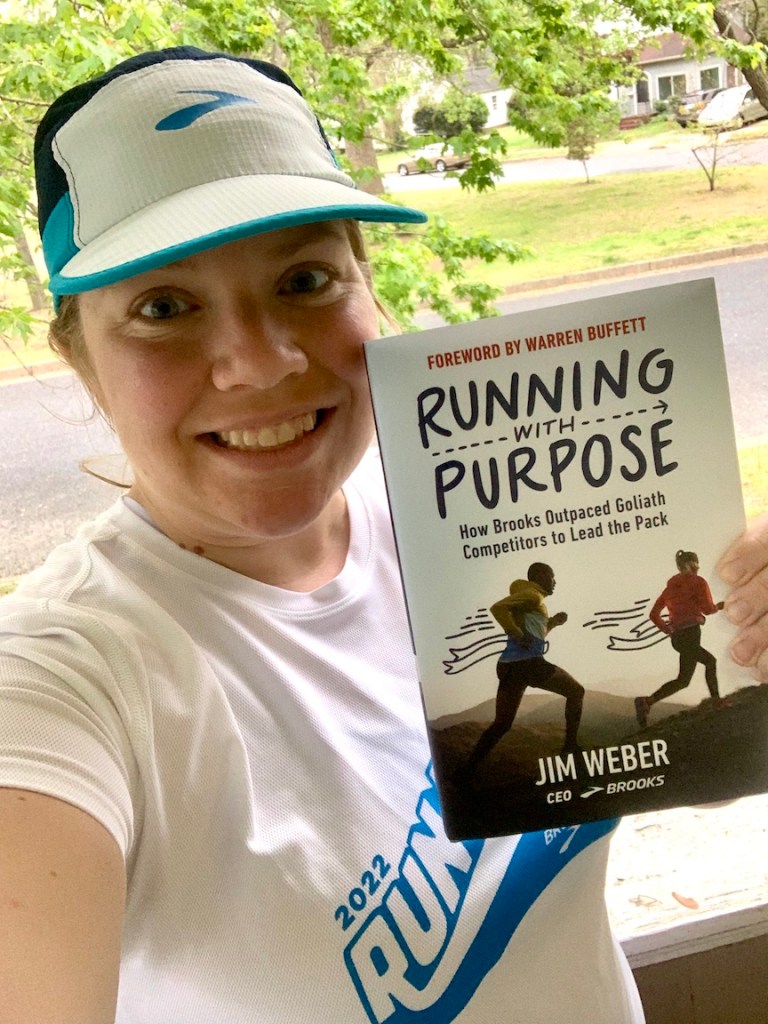 Vanessa Junkin, wearing a Brooks hat and Run Happy Team shirt, holds a copy of "Running with Purpose" by Jim Weber.