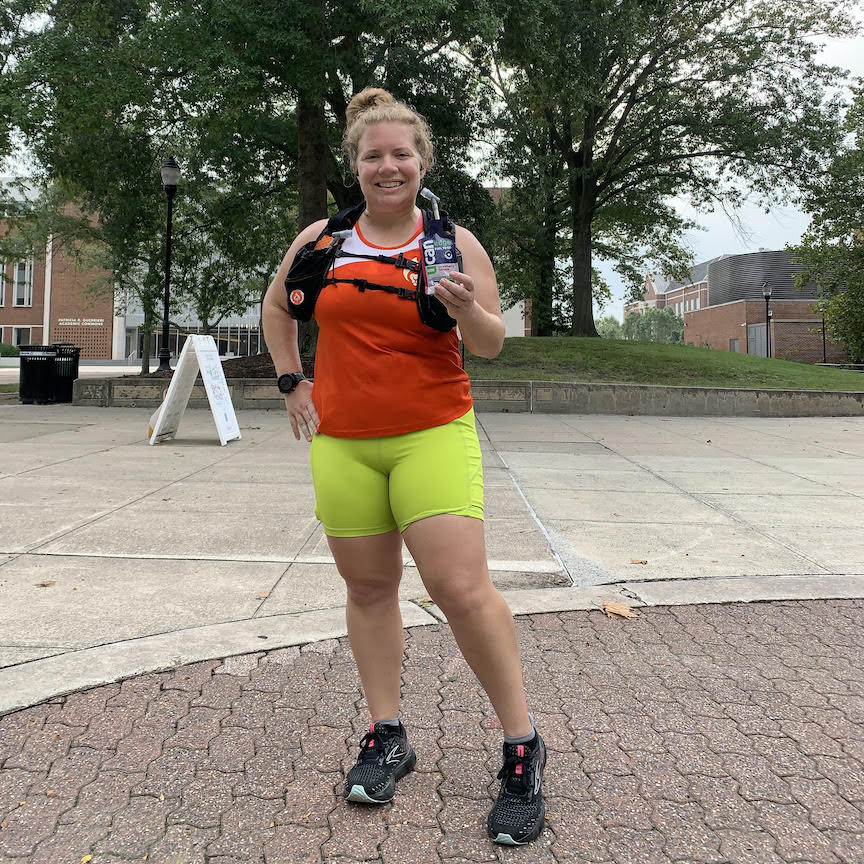 Vanessa Junkin stands in an orange BibRave tank and neon yellow shorts, holding a UCAN Strawberry Banana Edge gel. 
