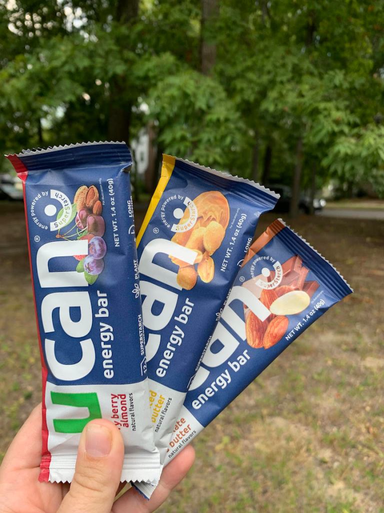 A hand holds three UCAN Energy Bars — Cherry Berry Almond, Salted Peanut Butter and Chocolate Almond Butter. 