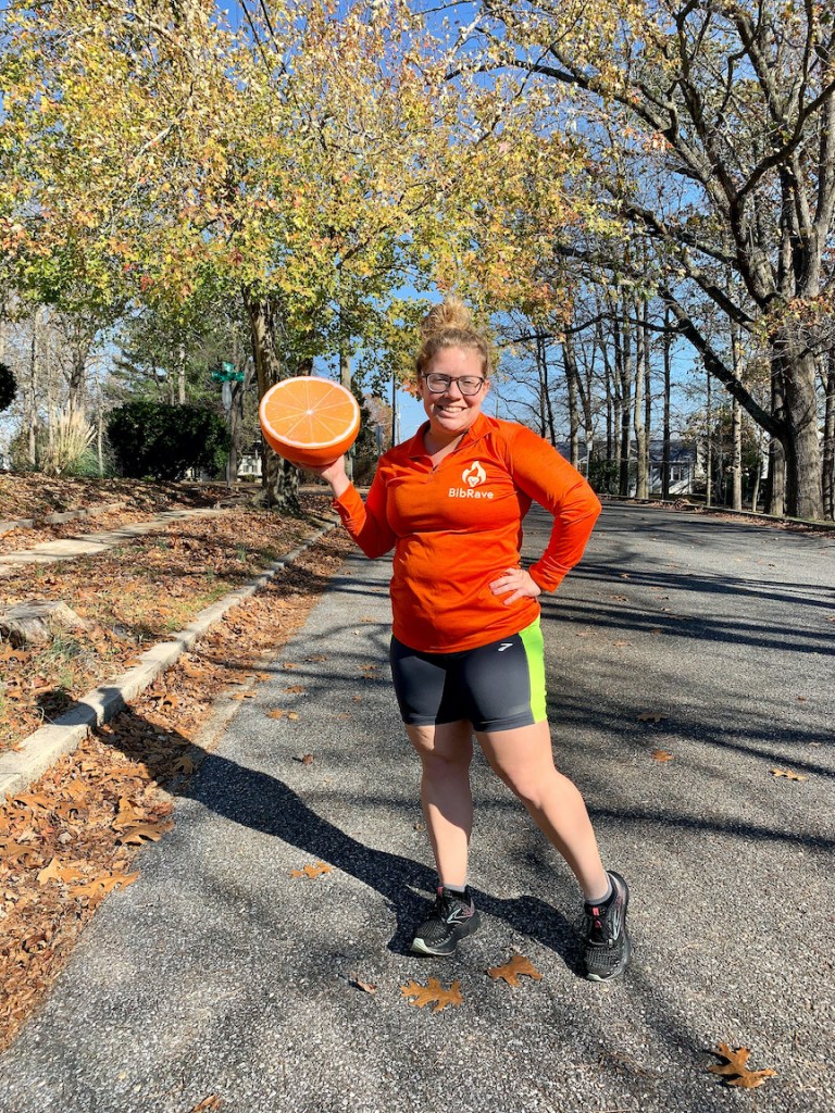 Vanessa Junkin, in an orange long-sleeved shirt and short, holds a large orange. 
