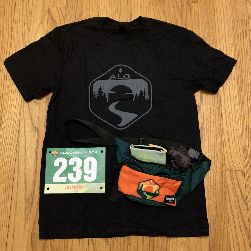 Swag set out on a wood floor — black T-shirt with logo, race bib and fanny pack with koozie and magnet in it. 