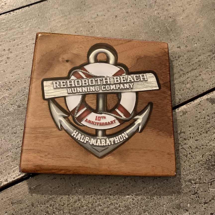 Wooden coaster with anchor-shaped medal inside. The medal has a life preserver that reads "Rehoboth Beach Running Company," "10th Anniversary" on the life preserver and "Half Marathon."
