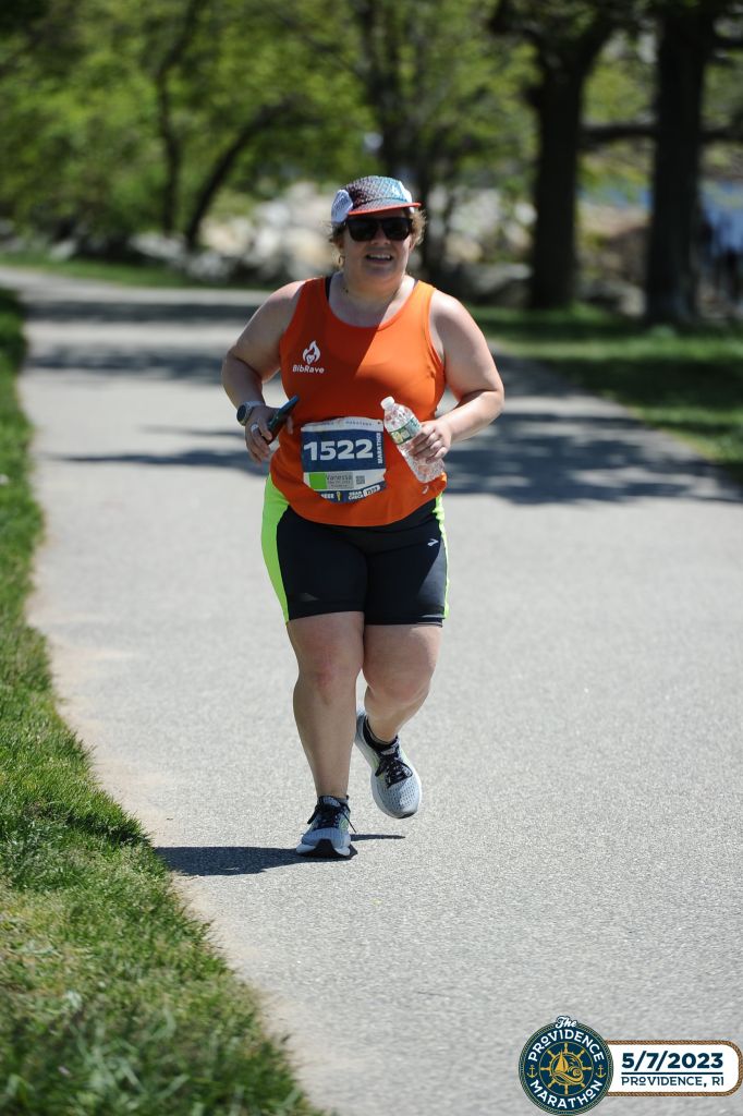 Female runner in a tank top and shorts runs on a paved trail with trees in the background. 