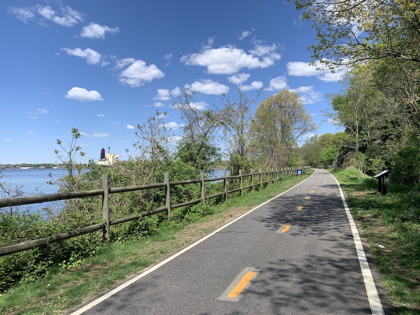 View of a bike path with a body of water to the left, a fence to the left, and trees on both sides. 