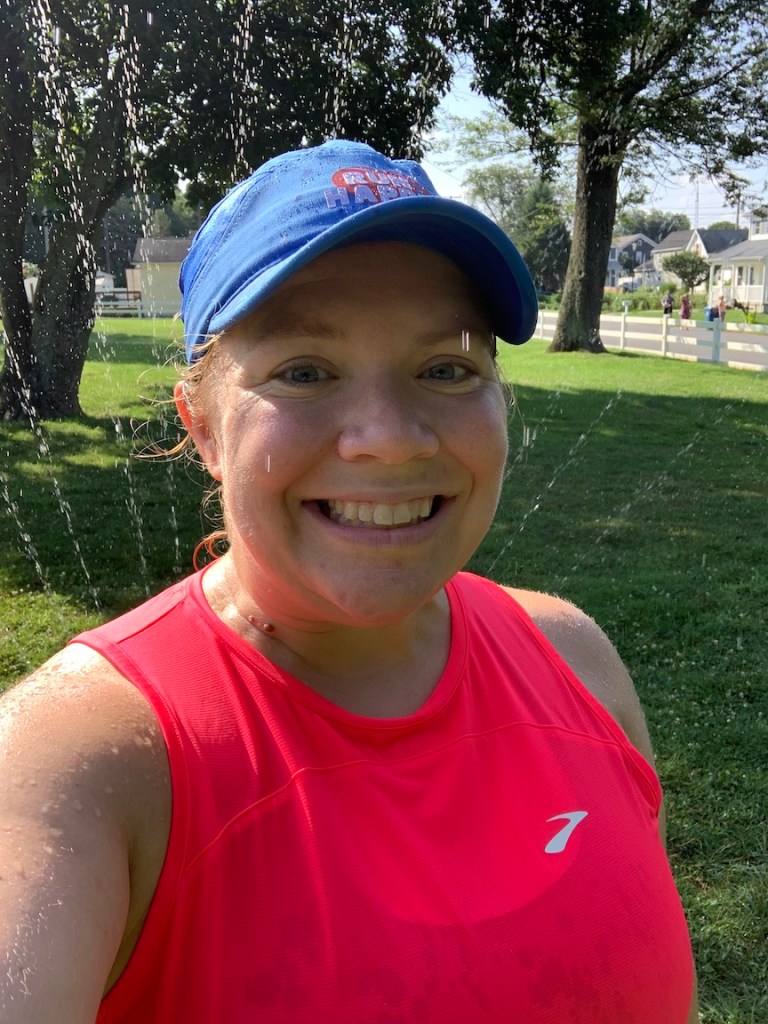 Selfie of female runner with the water from a sprinkler behind her.