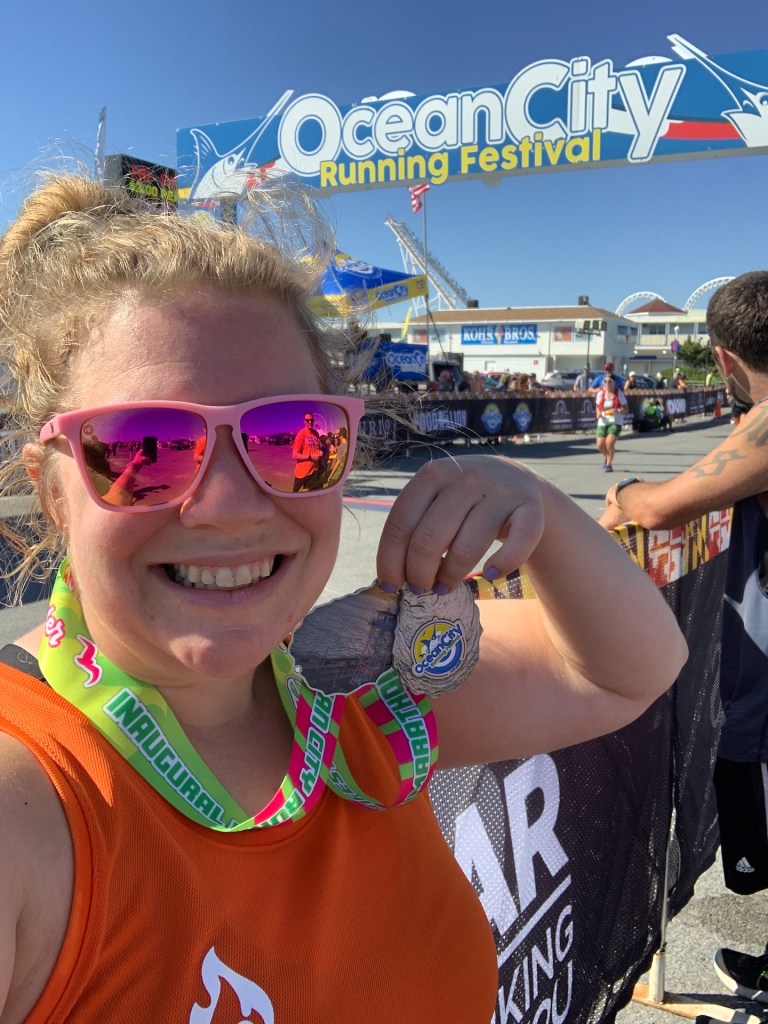 Selfie of Vanessa holding an oyster shell finisher medal, with the Ocean City Running Festival finish line in the background. 