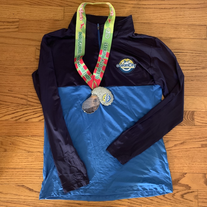 Blue 1/4 zip with the Ocean City Running Festival logo shown on the floor with the finisher medal. 