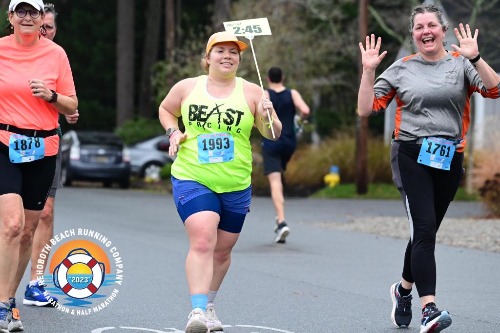 Female running pacer, holding a 2:45 sign, has a female runner on either side of her, one with her hands up. 