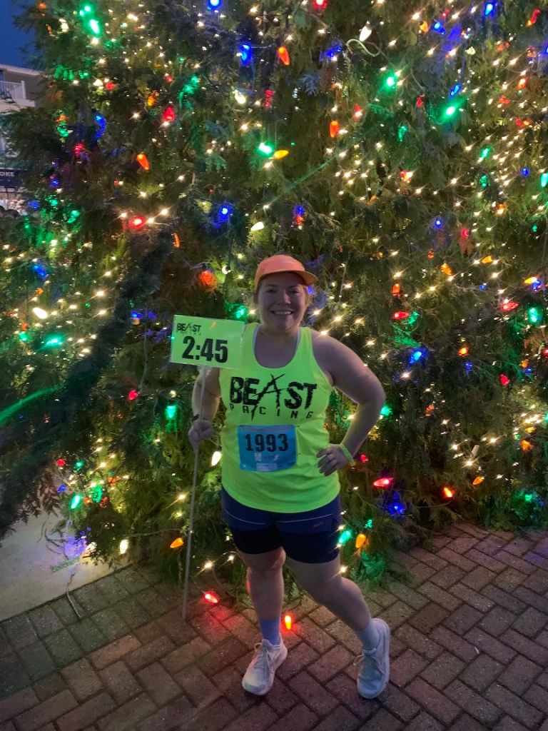 Vanessa Junkin, in Beast Pacing tank and shorts, holds a 2:45 sign and poses in front of a Christmas tree.