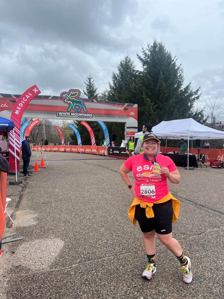 Photo of Vanessa Junkin posing by the REVEL White Mountains finish line with a finisher medal in hand. 